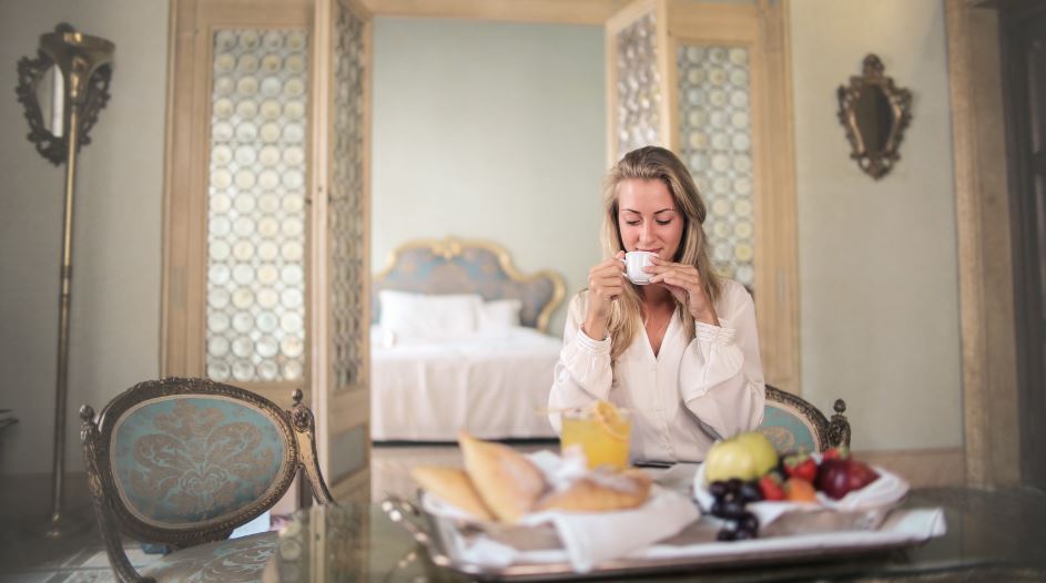 The Holistic Approach to Hospitality: A woman with blond hair drinks a cup of coffee in a hotel room. A breakfast is ready on the table and in the background is a made bed in soft blue and beige tones.