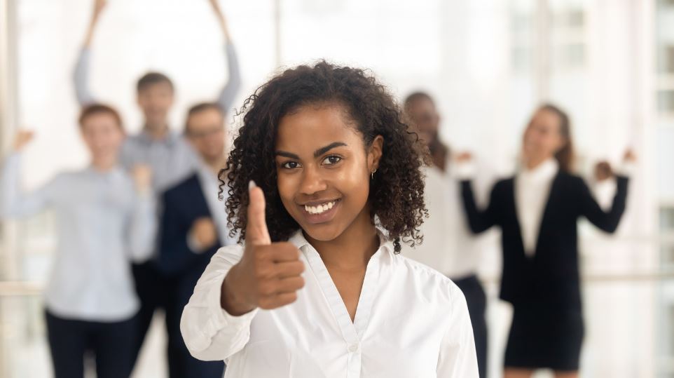 The Hostmanship Imperative Dark-skinned smiling young woman gives her thumbs up. With a somewhat blurred view in the background, happy employees with hands in the air.