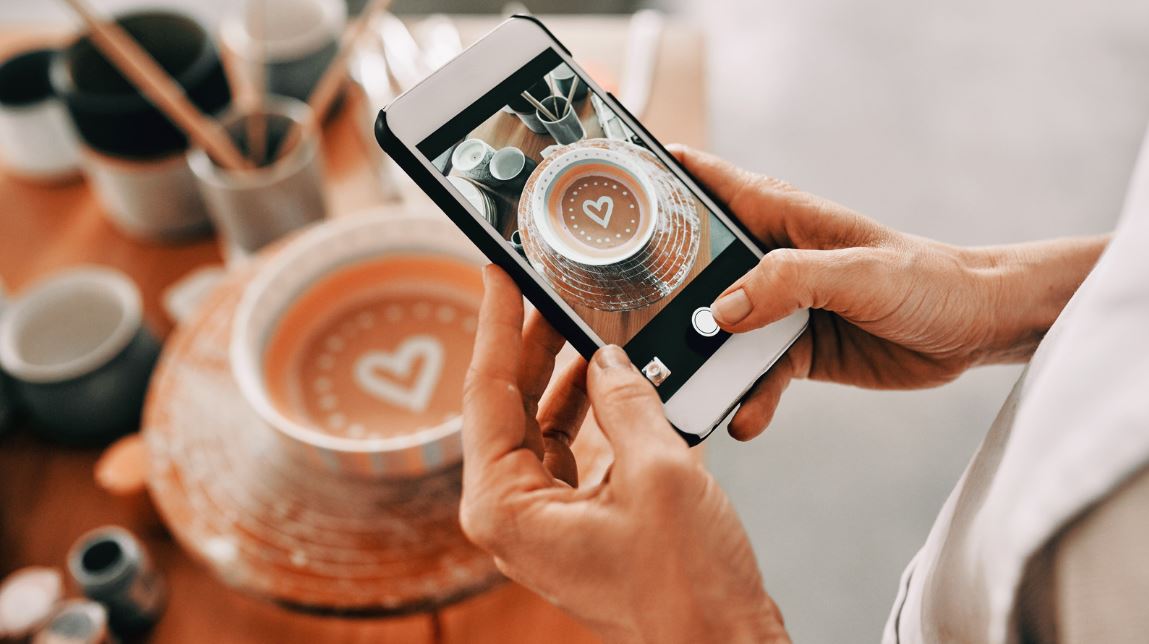 Go Digital but Stay Human: photo of a man's hands holding an iPhone takes a photo of a cup of coffee with a milk heart in the center. You see a real cup of coffee and an image of a photo on the iPhone.