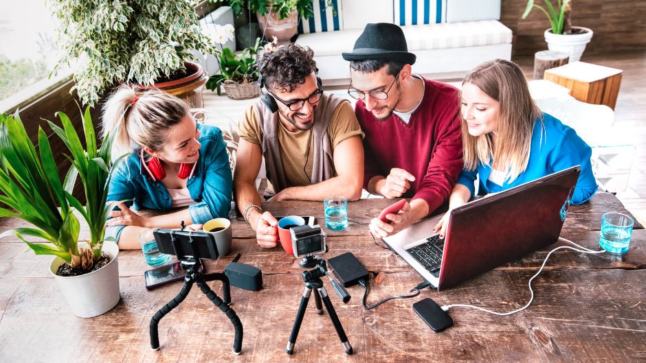 Rediscovering True Hospitality - 2 men and 2 women from Generation Z sit at the table in front of laptops, iPhones, headphones with a cup of coffee and talk to each other while laughing Inspiring Generation Z in the Digital Age -