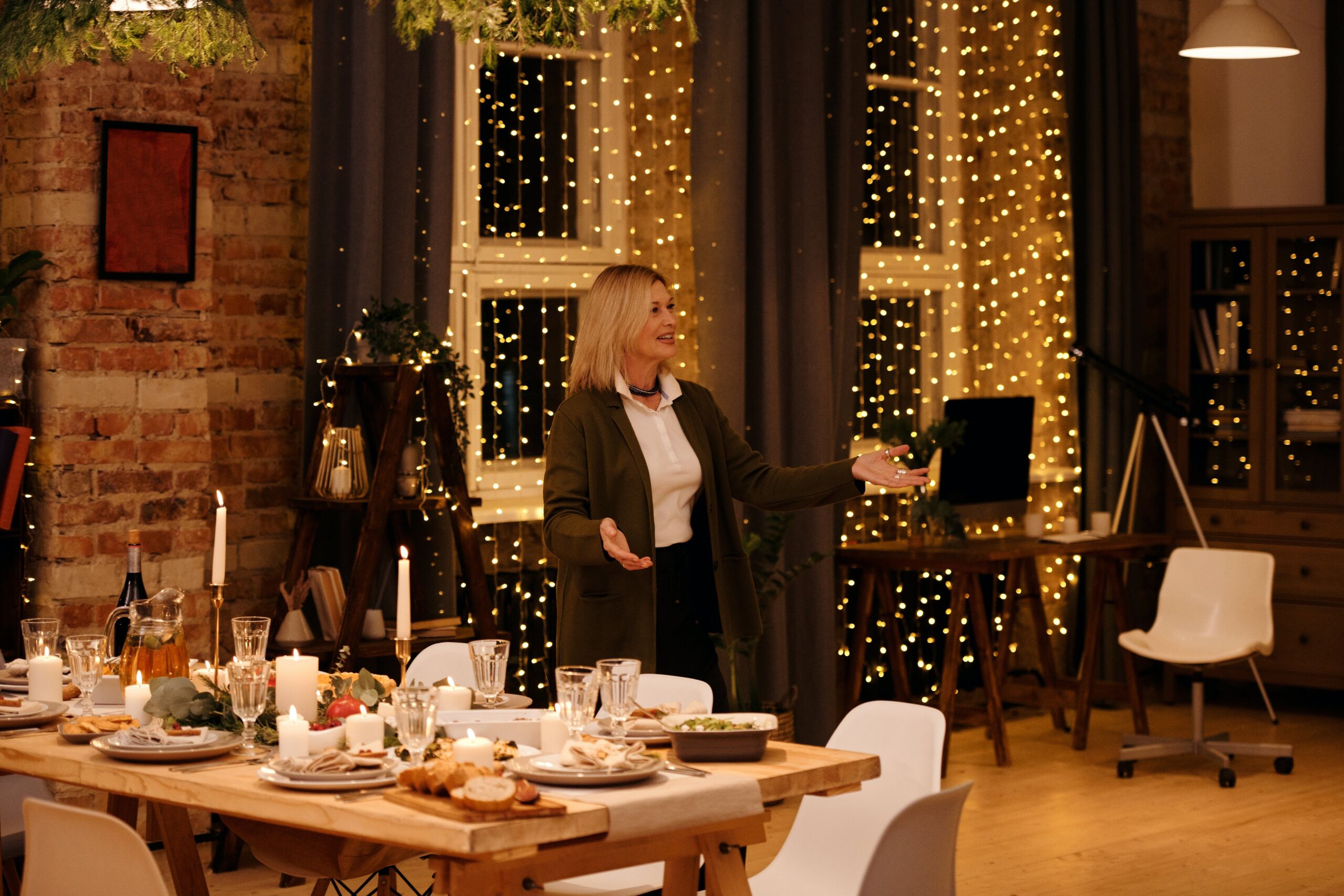 True Hospitality Makes Solid Happiness - Middle-aged lady receives her guests with open arms at an attractively set table. Room decorated with Christmas lights at the back.