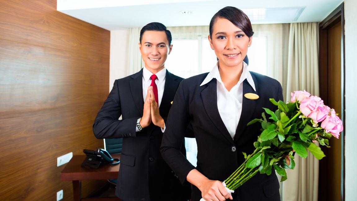 Unlocking Hospitality Chief Hospitality receptionist with bunch of roses and receptionist with welcome gesture, hands together, welcome guests